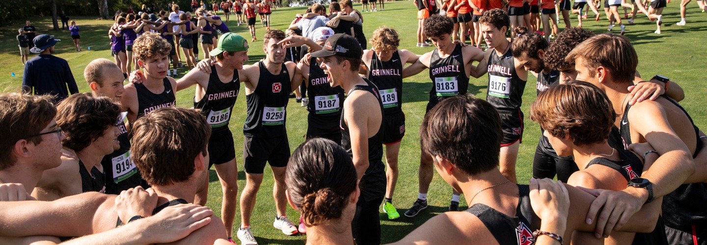 Grinnell men's cross-country team at a meet stands in a huddle with arms around each other as one stands in the center shouting other teams are visible in the background
